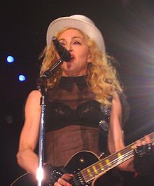 Picture of a middle-aged blond woman uptill the waist, singing in front of a microphone. Her hair is in waves and falls up to her shoulders. She appears to be wearing a black bra covered with a sleeveless netted covering and wears a white hat on her head. There are black gloves on her hand and she plays an electric guitar.