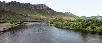 A dark river flowing between bushes and open ground, curving to avoid a high ridge