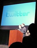 long view of stage with Stone and Dorsey speaking in front of a slide presentation