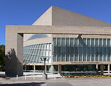 The Morton H. Meyerson Symphony Center in Dallas, a beige cube rising at an angle around a half-cone made of glass and steel. In front, a square archway overlooks a stone courtyard.