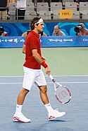 A dark-haired man is in a red shirt with white shorts and shoes and bandanna, which he is carrying his tennis racket in his right hand pointing towards the ground