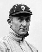 A man, wearing a baseball cap with the Detroit Tigers' Old English "D" logo in the center and an off-white baseball uniform with obscured lettering, faces forward towards the right.