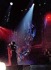 Image of a brunette woman in a black-and-red kimono moving to the right of a stage with red-lit backgrounds and a metallic tree like structure in the middle. A single source of light falls on her.