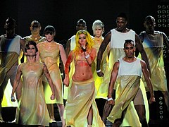 A group of dancers on stage, wearing light yellow colored dresses. In their middle is a blond singer wearing the same dress, but with blotches of red color all over her body