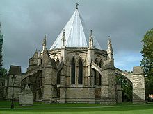 An external view of the Chapter House at Lincoln against a dark cloudy sky. The polygonal building has a steeply pointed lead roof, paired pointed windows and huge flying buttresses with splay out all around the building like spider's legs.