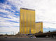 Ground-level view of a V-shaped, 45-story building with a golden, all-glass facade; a palm tree is visible in the foreground, and it partially blocks the view of the skyscraper.