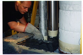 Construction of a test sample consisting of a mock-up concrete floor frame, complete with penetrants. The concrete frame measures approximately 5’ x 9’ x 4" (ca. 1.5 m x 2.3 m x 10 cm). It has a large hole in the centre with many traversing mechanical and electrical services. The penetrants extend 1’ (30 cm) into the furnace and 3’ (91 cm) on the unexposed side. A firestop mortar is being applied here. Notice the intumescent wrap strip surrounding the fibreglass pipe insulation. When the fire starts, this embedded intumescent swells to take the place of the melting insulation. The test was conducted in accordance with the Canadian firestop test method ULC in Scarborough, Ontario.[4][full citation needed]