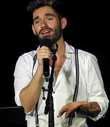 A brown-haired man with a beard and multiple tattoos performing on stage in a white shirt. She is holding a black microphone.