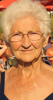 Close-up portrait photo of the upper chest, neck, and face of Johanna Quaas, a tan Caucasian woman with short styled white hair, glasses, smiling, and who is slightly squinting because the sun is in her eyes; she is wearing a black one piece bathing suit of which the top part is visible