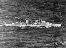 Black-and-white aerial photograph of a two-funnelled cruiser underway. Alternating light and dark bands are painted on the side of the ship.