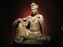 A wooden carving of a female human figure, sitting with one knee on the ground and one knee pointing up, with a hand resting on that knee. The carving includes loose fitting clothing, which is covered mainly in gold foil.