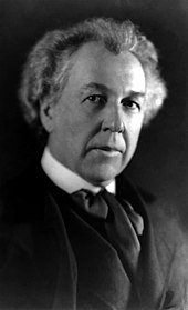 A portrait photograph of a middle-aged man with bushy white hair, wearing a dark suit