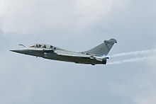 Pale gray jet aircraft flying towards left of camera. Smoke trial the aircraft's wing tips