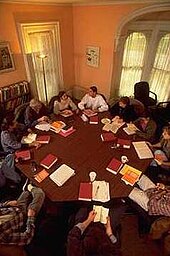 eleven people sitting around octagonal wood table with red books in front of them seen from above