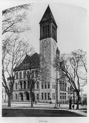 A light building with dark trim stands on a street corner; it has an arched entrance at left, a double-peaked roof, and a 200-foot tower at the closest corner.