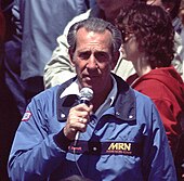 Ned Jarrett in broadcasting overalls and holding a microphone in his right hand