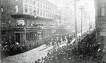 city street view from above, street seen diagonally from lower left to upper right, crowds of people line the street, horses, carriages and people march along it