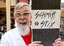 A photo of Paul Schmitt, with wood framed glasses, a white beard, and a white lab coat, holding a Schmitt Stixx Skateboard on his left side, and a big smile on his face.