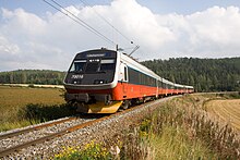 A four-car multiple unit running down a single track with fields on both sides and a forest in the background