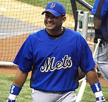 A man, wearing a blue baseball cap with an interlocking "NY" at the centre, a blue baseball uniform with the words METS across and a blue wristband around both wrists, smiles as he takes batting practice