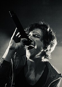 A shoulders-up portrait of Ricky Montgomery in concert. He is wearing a tank top and light raincoat, and is singing passionately into a microphone.