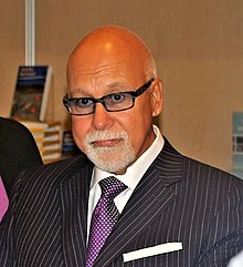 Photograph of René Angélil. Depicting his head an upper torso, the man has a bald head, tanned skin, and grey goatee beard and moustache. He is wearing spectacles, white shirt with purple necktie, and a pinstriped suit jacket.