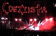 A view of the stage being filmed, with "COEXISTA" spelled out on the video screen, using an Islamic crescent as a "C", a Star of David as an "X", and a Christian cross at a "T". A camera in the middle shows the concert being filmed.