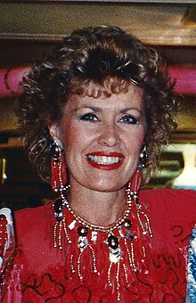 A head shot of a blonde woman in a red dress and a gold necklace