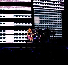 The picture is from a distance and shows a blond woman performing on a stage. She appears to wear red shorts and plays a pink guitar. Behind her, there are video screens showing horizontal graphical patterns