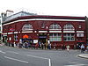 A red-bricked building with a rectangular, blue sign reading "HAMPSTEAD STATION" in white letters and people in front all under a white sky