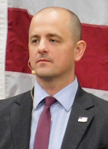 A man wearing a suit and a headset microphone, in front of the red and white stripes of a U.S. flag