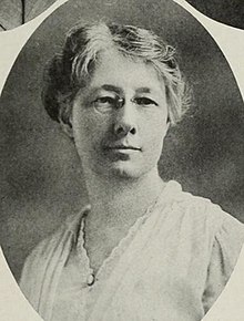 A middle-aged white woman, wearing eyeglasses and a white blouse. Her grey hair is dressed up off her neck and shoulders.