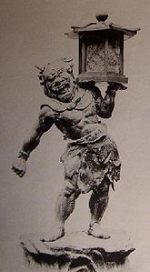 Front view of a stocky statue with a demon face. He is carrying a lantern on is left shoulder supported by his left hand. Black and white photograph.