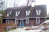 A brown house with low sloping black roof and dormer windows. There is a light snow covering on the brick and stone fountain and front lawn.