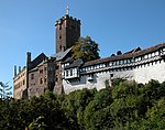 A castle perched along the edge of a wooded hill. The castle has grown in several stages and consists of sections in dark stone, lighter stone, white plaster and half-timber.