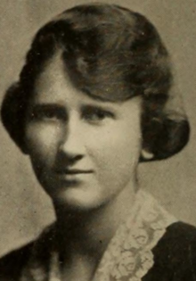 Young white woman with dark hair dressed in swept bangs, wearing a lace collar on a dark jacket