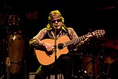 A man sitting, holding a guitar, with a microphone in front of him.