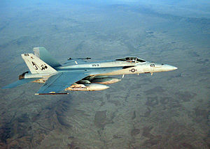 Top view of gray jet fighter banking away from camera. Under the fuselage is an external fuel tank