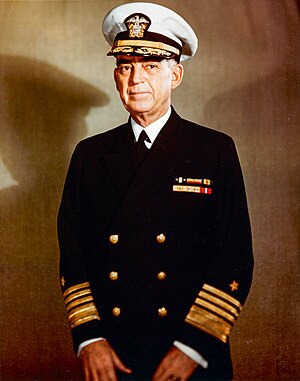 Man in dark blue suit and tie, wearing peaked cap and two rows of ribbons.