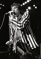 A man stands onstage with a guitar strapped across his chest. It hangs limp and unused as he focuses on singing into a microphone directly in front of him, which he grasps with his left hand. He wears a black-and-white, vertically striped suit.