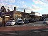 A brown-bricked building with three brown-bricked chimneys, some scaffolding on the side, and seven vehicles in the parking lot in front