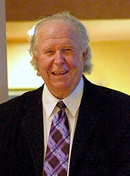 A man with disheveled light gray hair wearing a black suit jacket and purple striped tie smiles and looks directly ahead.