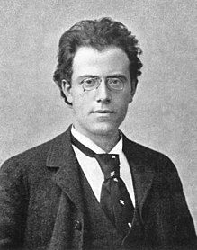 A young man wearing rimless spectacles looks straight out of the picture. He is formally dressed in dark jacket, white shirt and loose necktie. His longish hair is brushed back to reveal a wide forehead.