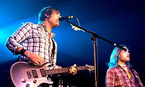 A man with a guitar singing into a microphone, with another musician playing a bass guitar