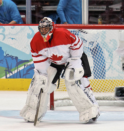 An ice hockey goaltender wearing a white and red jersey with white pads and a red mask. He is bent forwards with his hands at his knees and his head looking forward, eyes looking up.