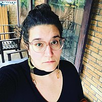 Carmen Maria Machado from the shoulders up. She is wearing glasses, hoop earrings, and a black top with a cutout neckline. She is looking at the camera.