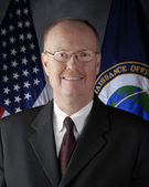 A Caucasian male posing for a professional photo wearing a black suit with a white dress shirt and burgundy tie. A United States flag is seen in the background on the left, while a flag with the globe logo of the National Reconnaissance Office can be seen on the right.