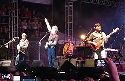 Four middle-aged men playing musical instruments on a stage in front of an audience