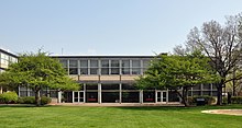 a low glass and steel building behind a sidewalk and lawn and trees