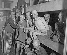 A group of malnutrished prisoners, some of them naked, some dressed in prisoner clothing, fill the interior of one of the barracks, as well as two lower levels of a three-storey bed.